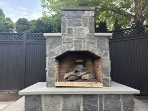 Stone masonry outdoor fireplace design with stacked stone and concrete base, surrounded by a wooden fence, showcasing luxury landscape features by Soft Wind Landscape & Masonry.
