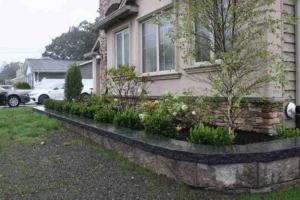 Landscape design featuring a stone masonry border with lush greenery and flowering plants surrounding a residential property in New York.