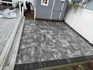 Newly installed masonry patio featuring textured gray pavers, adjacent to a wooden deck and shed, showcasing Soft Wind Landscape & Masonry's outdoor design expertise.
