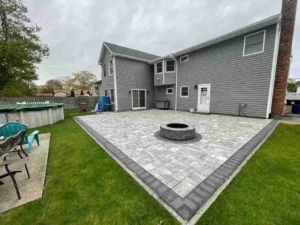 Backyard patio featuring a circular stone fire pit, surrounded by manicured grass and a modern home, showcasing landscape design by Soft Wind Landscape & Masonry in New York.